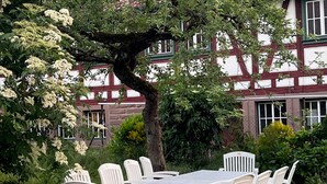 A round table with white chairs set outdoors, surrounded by greenery and a building with a red-tiled roof. - Kanne Cottage Flaire Suite with Stream and Meadow (Epfendorf/Trichtingen)