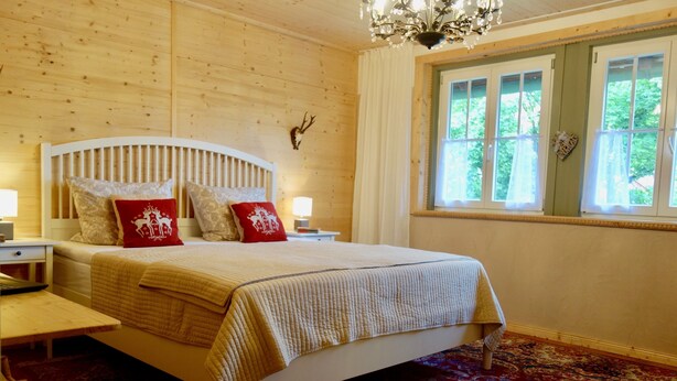 A bedroom with a wooden headboard, a bed with a quilt, two bedside tables with lamps, a chandelier, a window with curtains, and a deer head decoration on the wall. - Kanne Cottage Flaire Suite with Stream and Meadow (Epfendorf/Trichtingen)