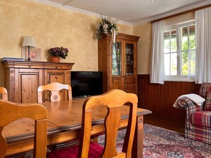 Une table de salle à manger en bois, des chaises, un téléviseur, un meuble avec des portes en verre et une fenêtre avec des rideaux.