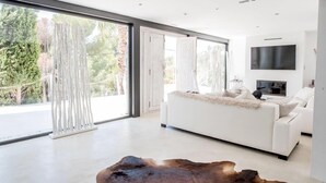 Ein modernes Wohnzimmer mit einem weißen Sofa, einem Flachbildfernseher und großen Fenstern mit Blick auf Bäume und ein Gebäude.