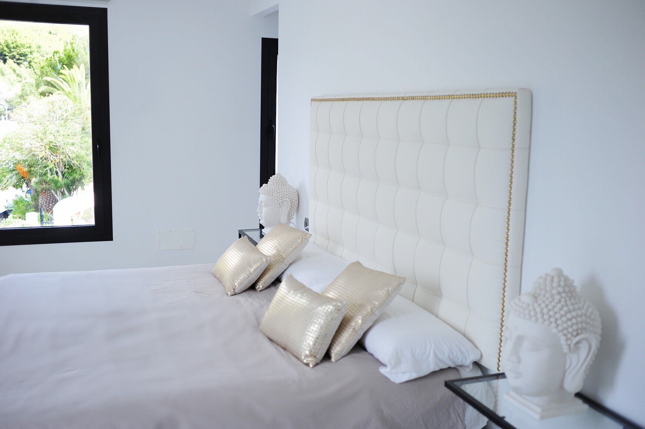 A bed with a tufted headboard, a window with a view of greenery, and a white bust sculpture on the nightstand.