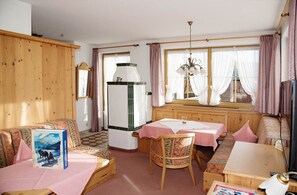 A cozy room with a wooden bench, a table with a pink tablecloth, a chair, a refrigerator, and a window with curtains. - Winklmoosalm Vacation Rental (Reit im Winkl)