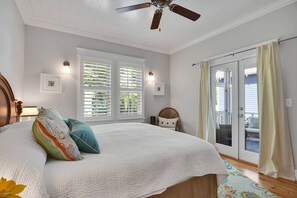 Quarto com uma cama grande, ventoinha no teto, duas janelas com persianas, uma cadeira e uma porta de correr para área externa.