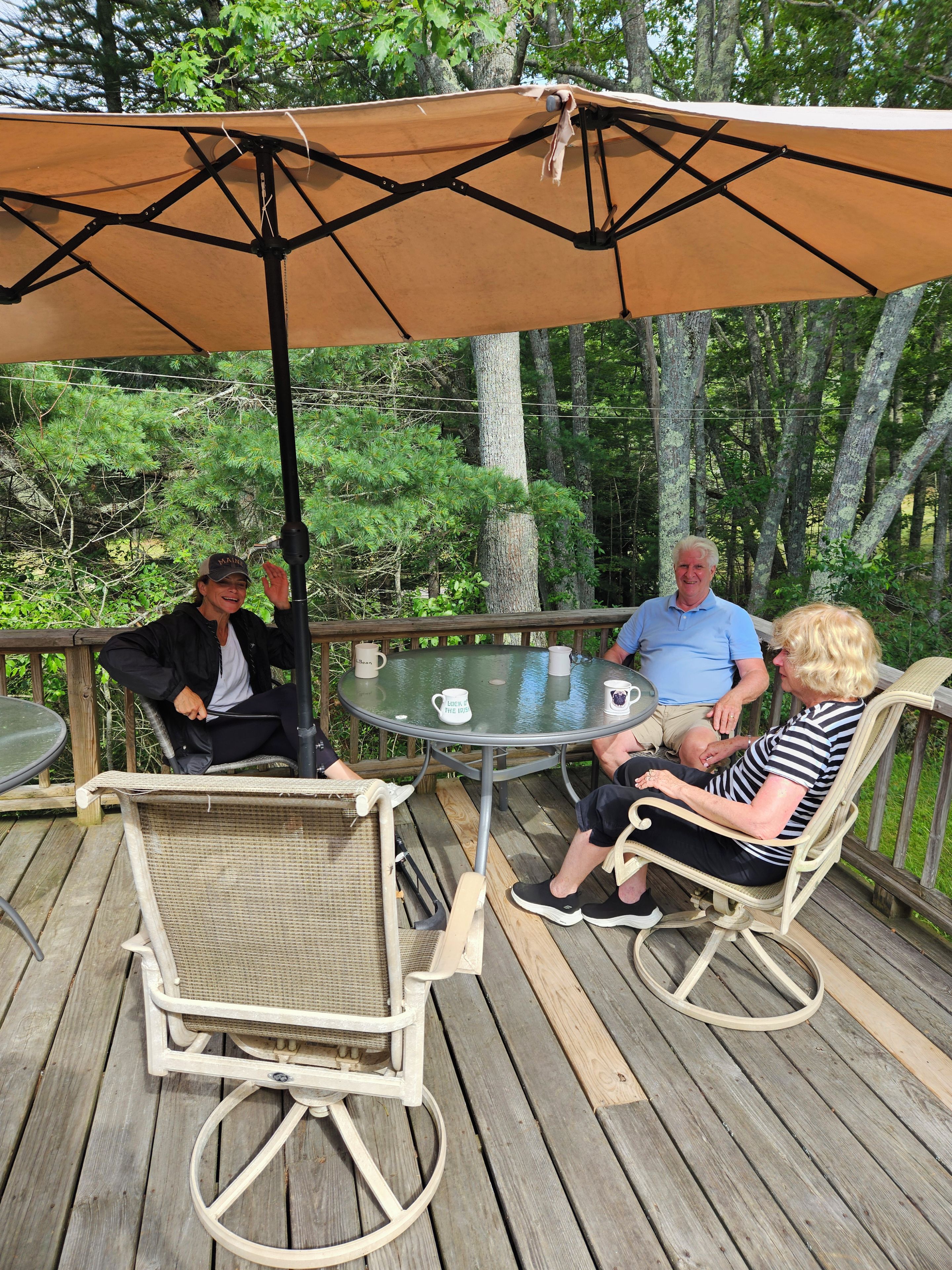 A wooden deck with a round glass table, wicker chairs, and a large umbrella providing shade.