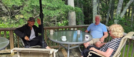 A wooden deck with a round glass table, wicker chairs, and a large umbrella providing shade.