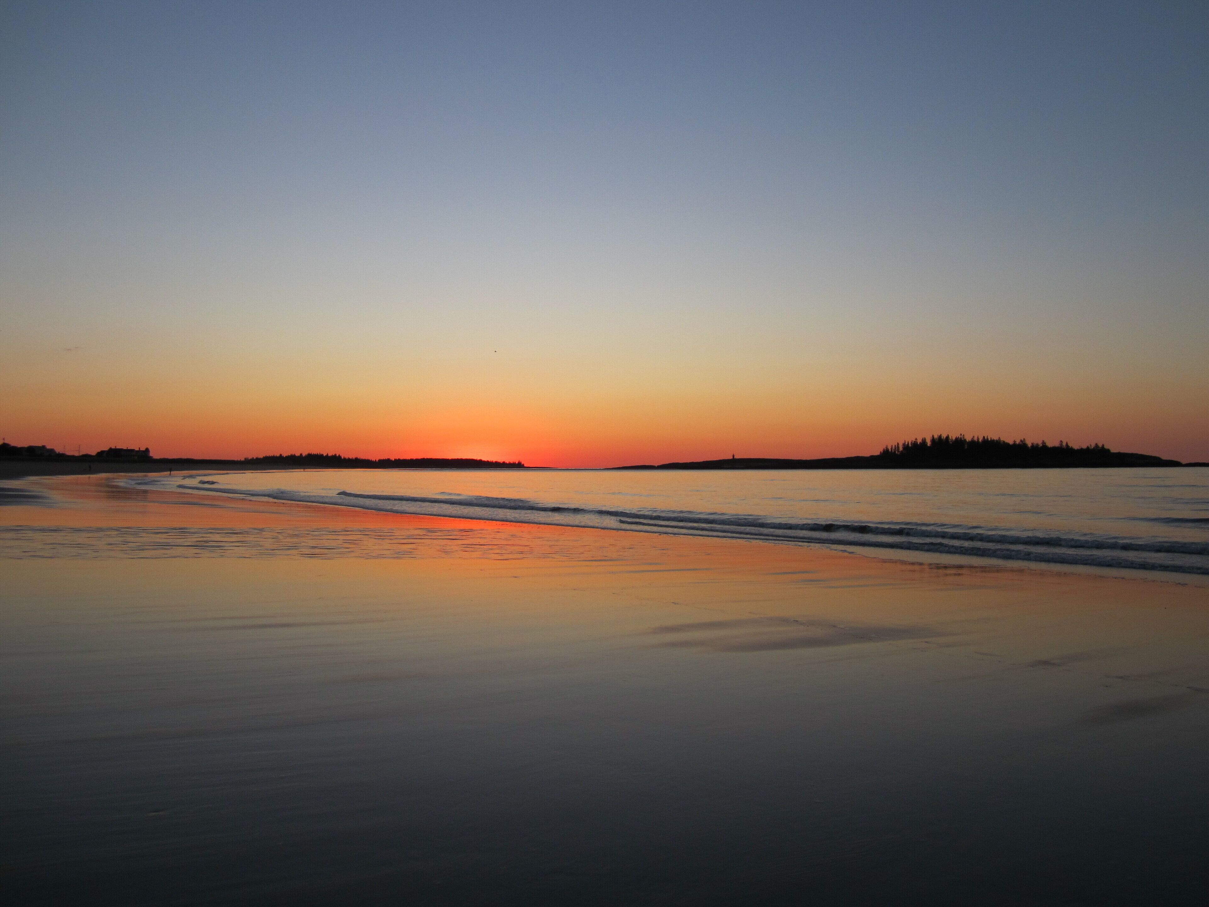A beach at sunset with calm waters and a distant shoreline.