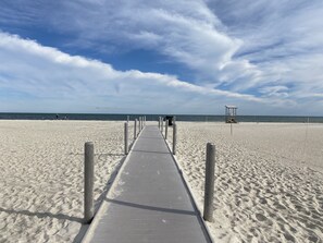 Een strand met een betonnen pad dat naar het water leidt, houten palen langs het pad, en een redershut in de verte.
