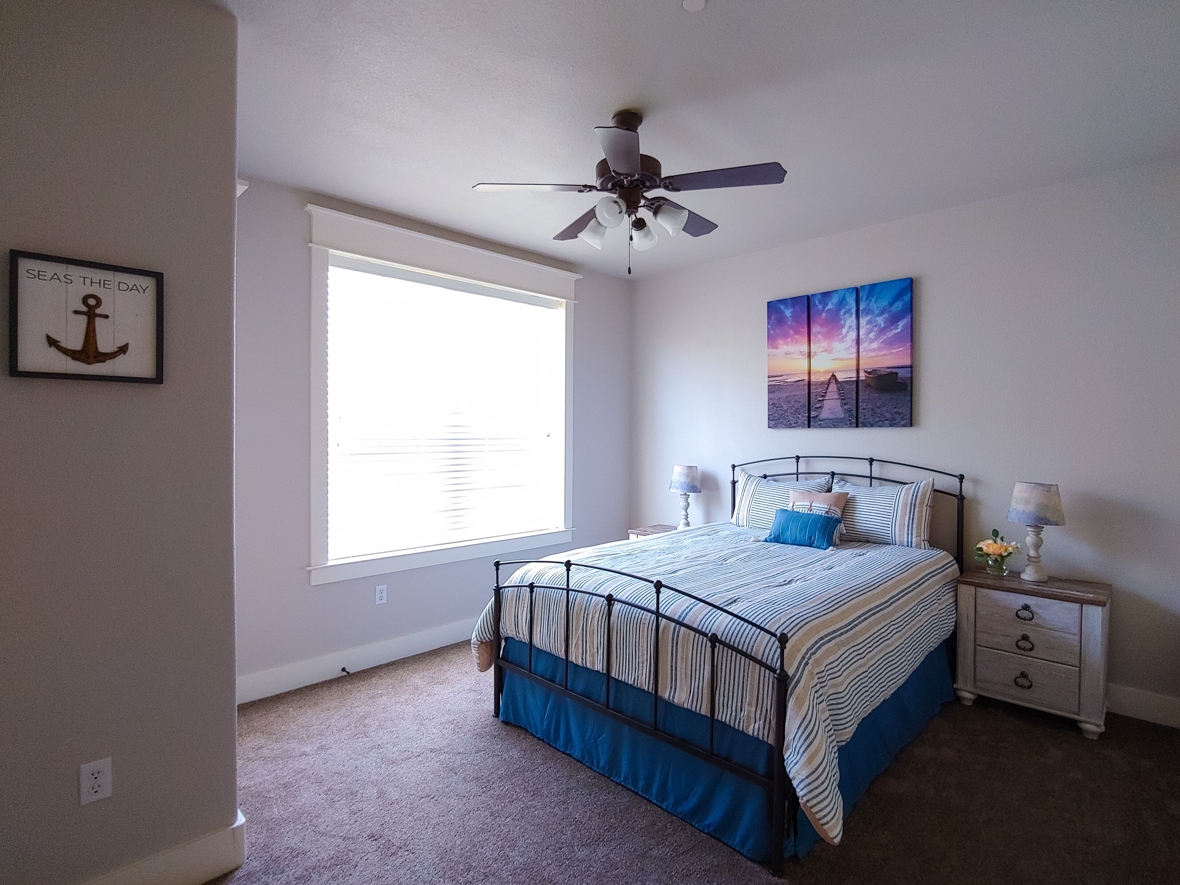 Une chambre à coucher comprenant un lit, un ventilateur de plafond, une fenêtre avec des stores, une table de chevet avec une lampe et un tableau accroché au mur.