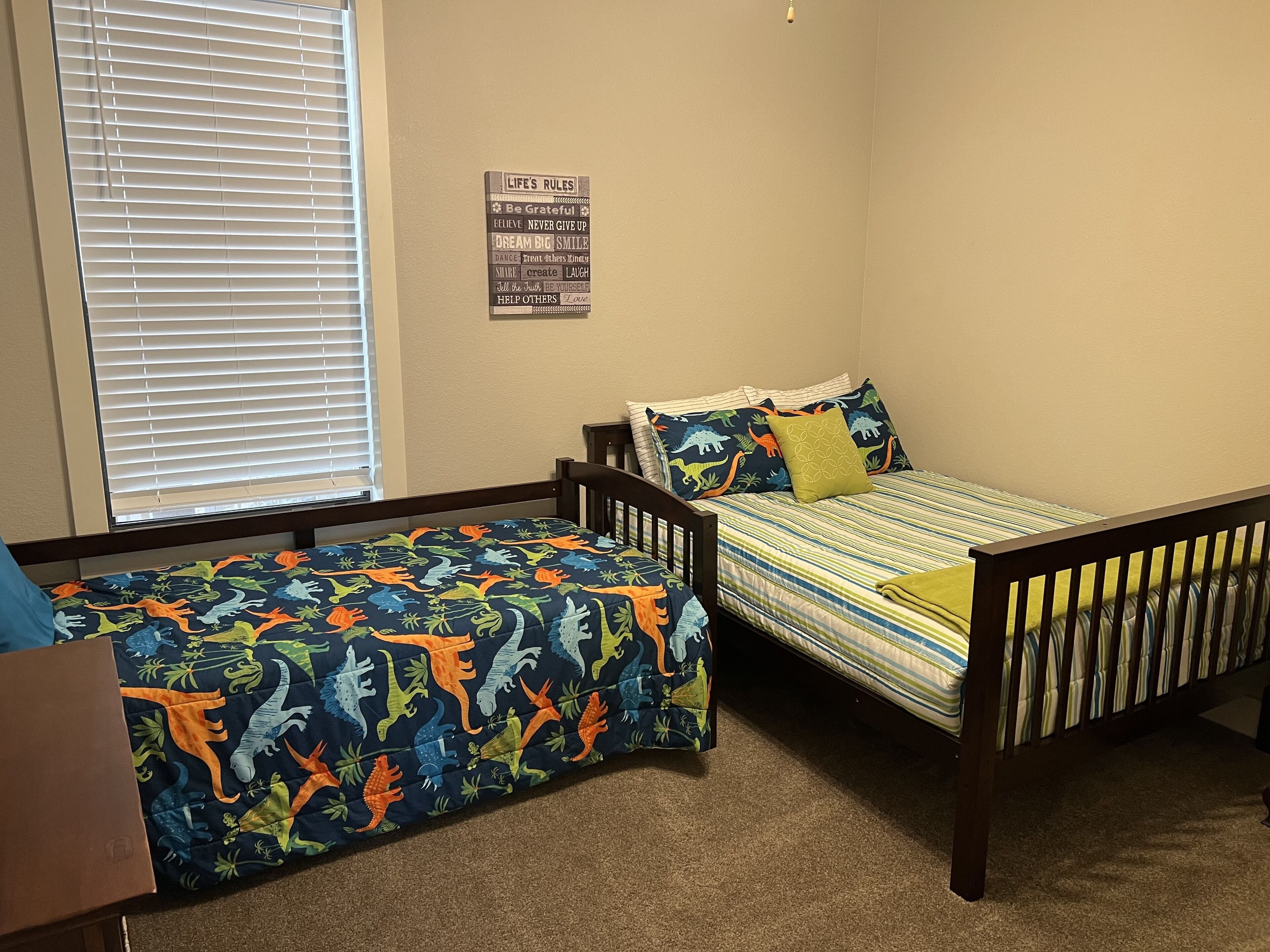 A bedroom with two single beds, a window with blinds, a wall-mounted sign with motivational phrases, and a carpeted floor.