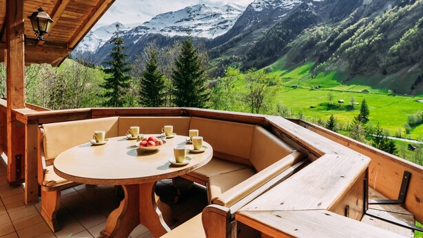 Um deck de madeira com uma mesa redonda e bancos, com vista para uma paisagem montanhosa com vales verdes e picos nevados.