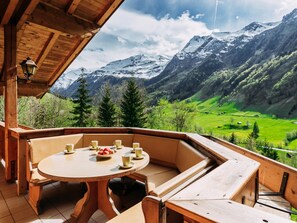 Una terraza de madera con una mesa redonda y bancos, con vistas a un paisaje montañoso con valles verdes y picos nevados.