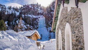 Um edifício de pedra com janelas em arco, uma cabana de madeira na neve e montanhas ao fundo.