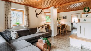Uma sala de estar aconchegante com sofá seccional, teto de madeira e área de cozinha ao fundo.