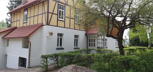 Ein traditionelles Fachwerkhäuschen mit weißer Fassade und roten Ziegeldach, umgeben von Grünpflanzen.