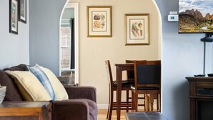 A living room with a sofa, a dining table with chairs, framed botanical prints on the wall, and a view of a mountainous landscape through a window. - Historic Drake Cottage 30 Day Rental. Across from Drake Park, Mirror Pond, downtown Bend. Furnished. (Bend)