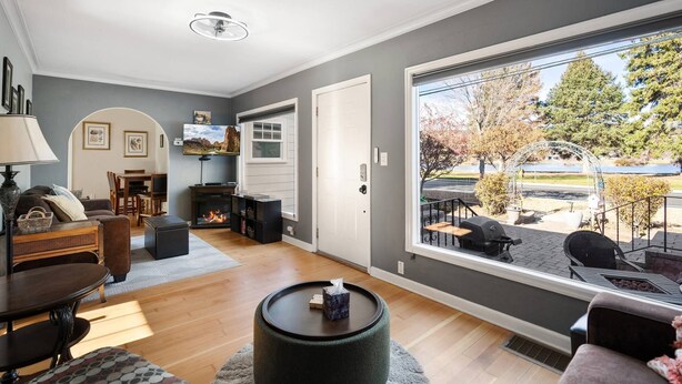 A living room with a fireplace, a television, and a large window overlooking a garden. - Historic Drake Cottage 30 Day Rental. Across from Drake Park, Mirror Pond, downtown Bend. Furnished. (Bend)