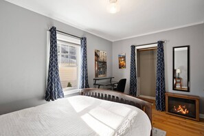 A bedroom with a bed, a desk, a chair, a fireplace, and a window with curtains. - Historic Drake Cottage 30 Day Rental. Across from Drake Park, Mirror Pond, downtown Bend. Furnished. (Bend)