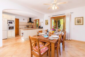 Une cuisine avec des meubles blancs, un four à micro-ondes et un four traditionnel. On y trouve une table à manger avec des chaises et un ventilateur de plafond.