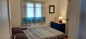 Ein Schlafzimmer mit Bett, Nachttisch mit Lampe, Kommode, Fenster mit Vorhängen und einem gerahmten Bild an der Wand.