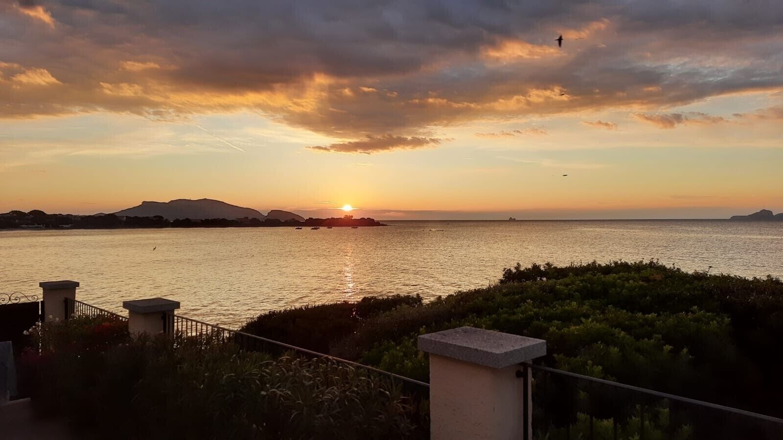 Panorama al tramonto su un corpo d'acqua con rilievi in lontananza e una ringhiera in primo piano.