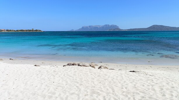 Uma praia com águas turquesa cristalinas, areia branca e formações rochosas ao longe.