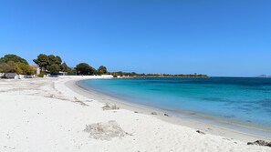 Ein Strand mit weißem Sand, klarem türkisblauem Wasser und einer fernen Küstenlinie mit Gebäuden und Bäumen.