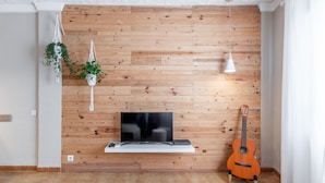 Een kamer met een houten wand, een televisie op een witte plank, een gitaar en hangende planten.