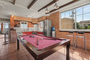 Una mesa de billar en una habitación con pared de ladrillo, un ventanal grande y una zona de cocina.