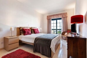 Un dormitorio con cama, mesitas de noche, cómoda, alfombra roja y ventana con cortinas.