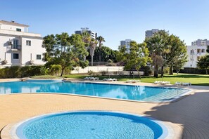 Una piscina con hidromasaje, rodeada por una terraza pavimentada, y edificios y árboles al fondo.