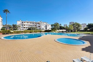 Un área de piscina con una terraza de madera, sillas de descanso y un edificio al fondo.
