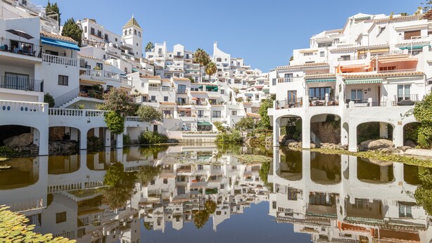 Une ville côtière aux bâtiments blancs, bordée d’une étendue d’eau calme et miroitante, et surplombée d’un ciel dégagé.