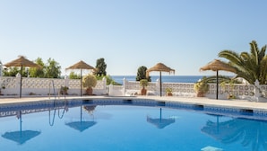 Une piscine d’un bleu éclatant, agrémentée d’une bordure décorative, entourée de clôtures blanches et de palmiers, avec une vue sur la mer en arrière-plan.