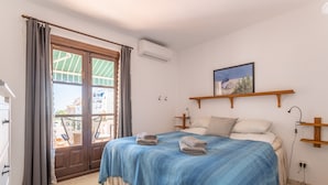 Une chambre avec un lit, une fenêtre avec des rideaux, un climatiseur fixé au mur, une étagère en bois et une vue sur l’extérieur.