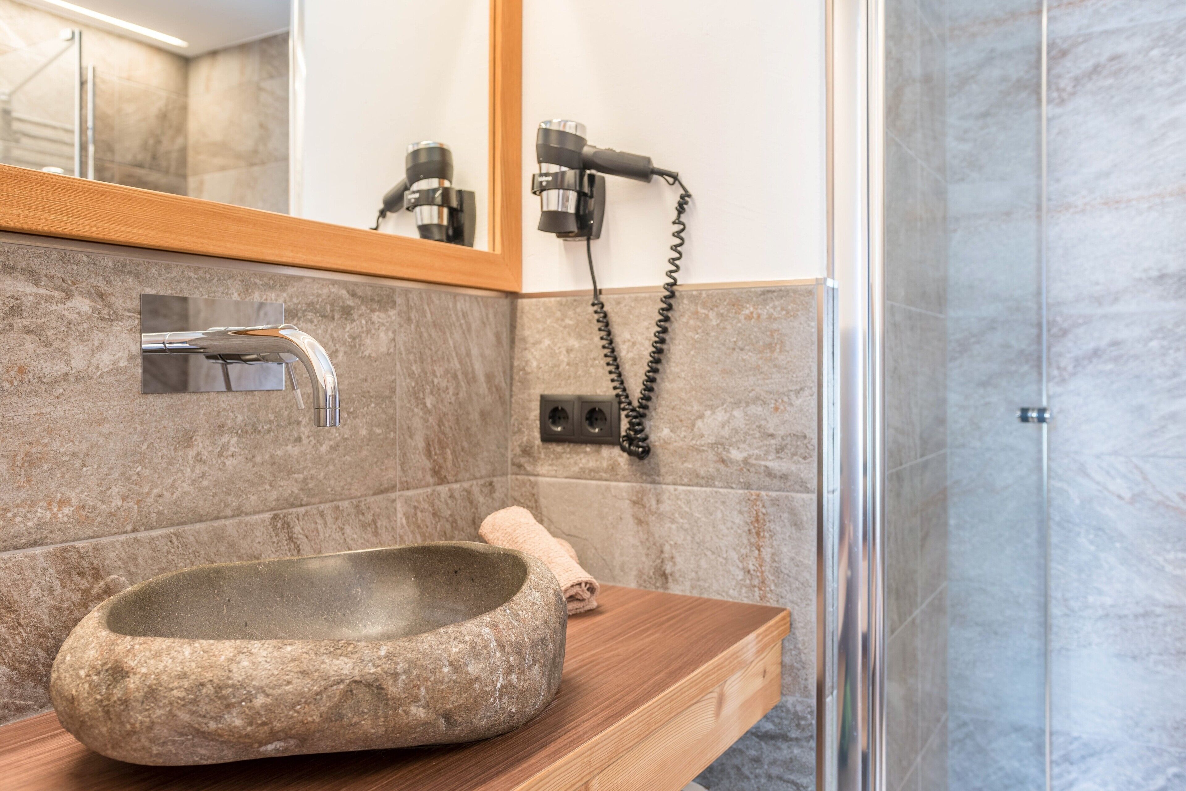 A bathroom with a stone sink, a wooden countertop, a mirror, a hairdryer, and a shower area.