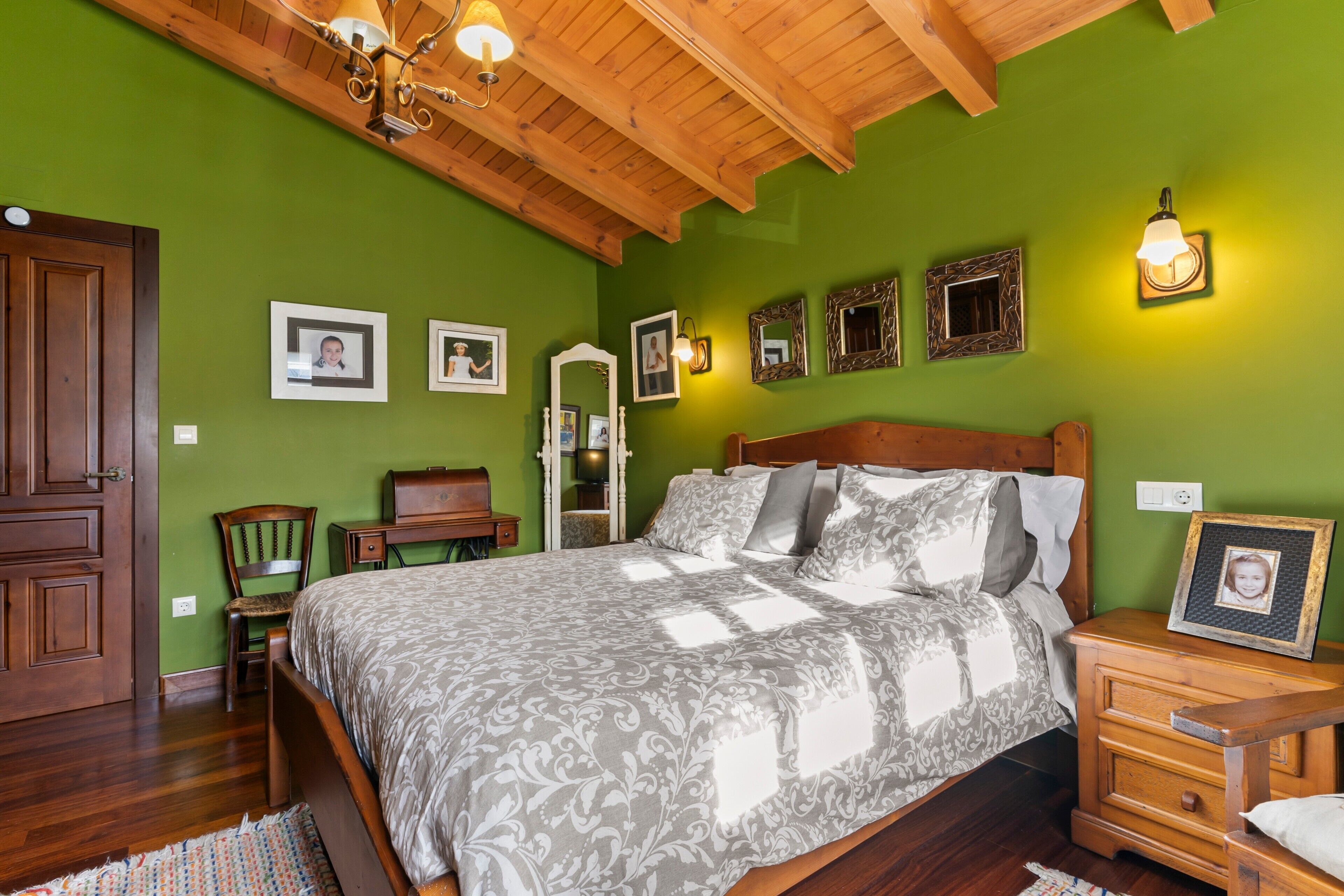 A bedroom with a wooden ceiling, a bed with patterned bedding, a nightstand, a chair, and framed pictures on the wall.