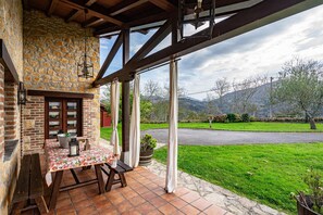 A covered patio with a stone wall, wooden beams, a table with a floral tablecloth, and a view of a green landscape and road. - Holiday Home La Milonga with Garden & Terrace (Piloña)