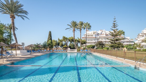 A swimming pool with clear blue water, surrounded by lounge chairs and palm trees, with a building in the background. - Spacious and Fantastic Views Apartment with Sea View, Pool, Garden & Wi-Fi (Nueva Andalucía)