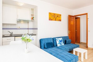 A modern kitchen with white cabinets, a white dining table with a vase, a blue sofa with a white table, and a wooden door.