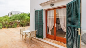 A patio with a table and chairs, a sliding glass door with curtains, and a white building with green shutters. - Felipe 1-4 Apartment with Terrace, Shared Pool & Wi-Fi (Tourist Taxes Included) (cala dor)