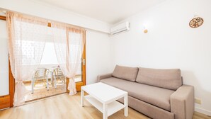 Un salon avec un canapé beige, une table basse blanche et une porte vitrée donnant sur un espace extérieur aménagé avec des chaises.