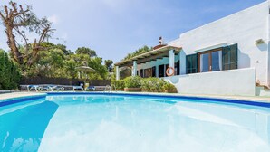 Une piscine à l’eau d’un bleu limpide, un espace extérieur aménagé et abrité, et un bâtiment à la façade blanche et aux volets bleus.