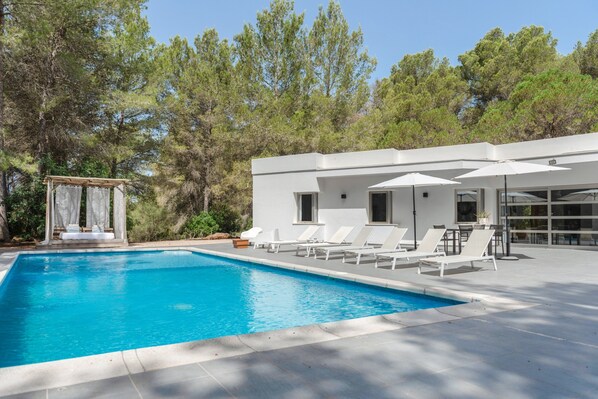 A swimming pool with lounge chairs and umbrellas, surrounded by trees and a modern building.