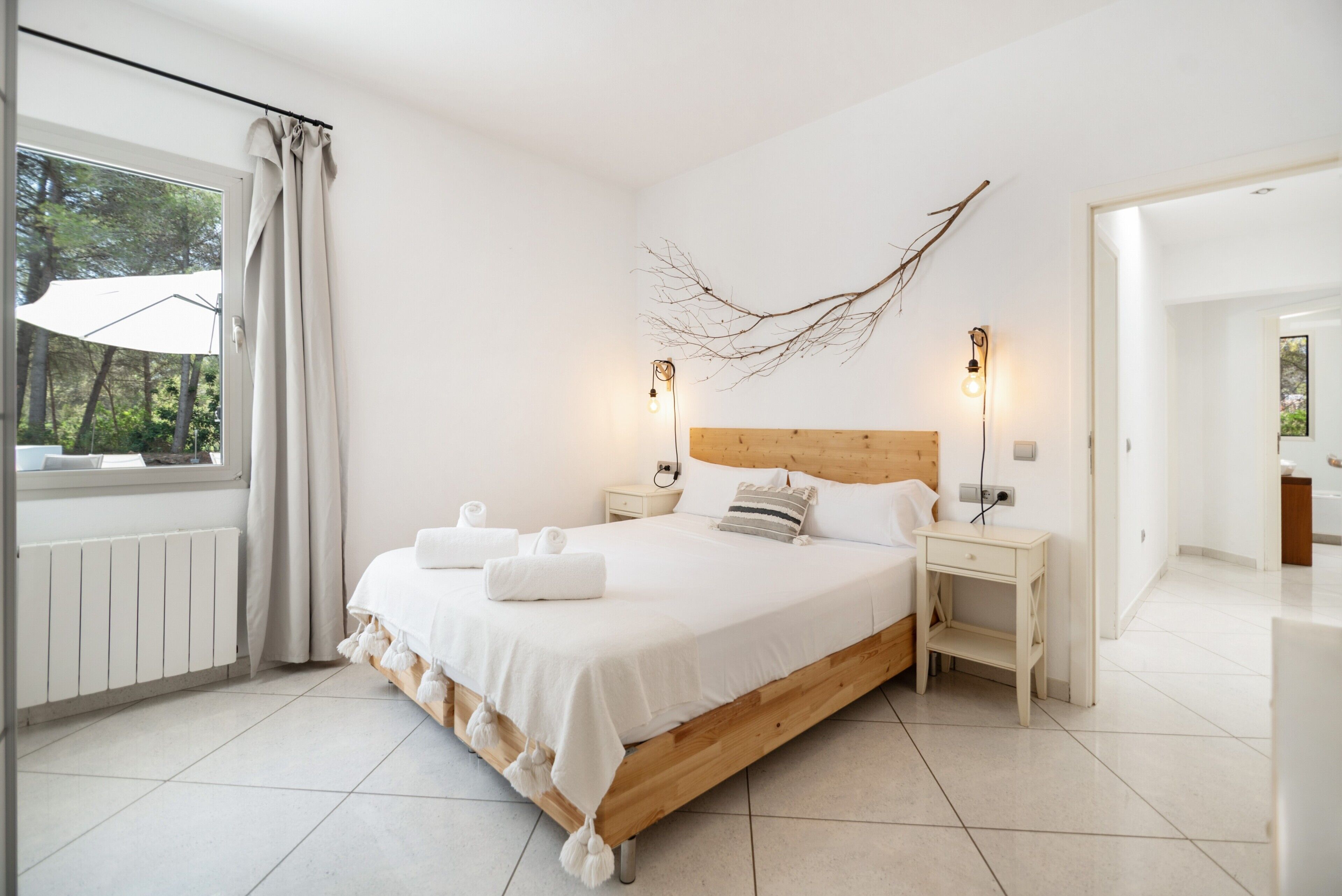 A bedroom with a wooden bed, white bedding, two bedside tables, a wall-mounted branch decoration, and a window with curtains.