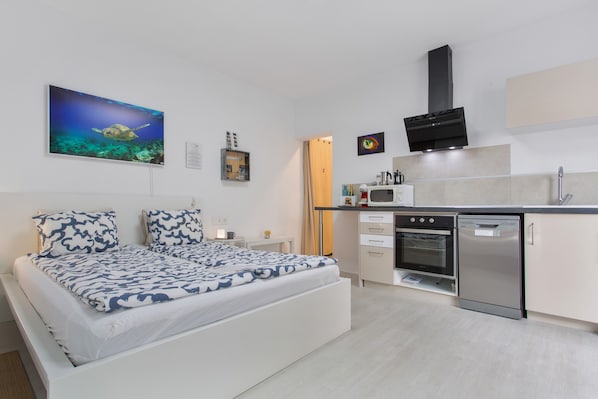 A bedroom with a bed, a kitchen with a stove, oven, and dishwasher, and a wall with a framed picture.