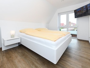 A modern bedroom with a white bed, a bedside table with a lamp, and a TV mounted on the wall.