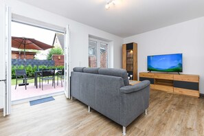 Un salon moderne avec un canapé gris, un meuble TV en bois et une porte-fenêtre coulissante donnant sur une terrasse extérieure.