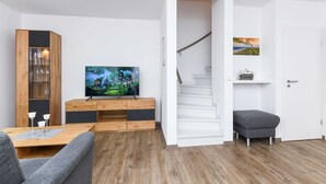 A modern living room with a wooden cabinet, a flat-screen TV, a staircase, and a grey ottoman.