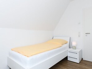 A single bed with a white frame and a yellow blanket, a bedside table with a lamp, and a white door. - Vacation Home in Meer, Germany (Dornum)