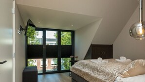 Un dormitorio moderno con una cama grande, un escritorio pequeño y vistas a un edificio a través de un amplio ventanal.